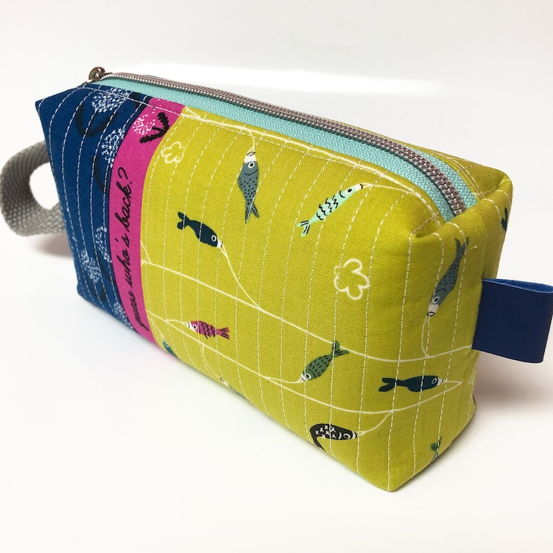 May include: A yellow fabric zippered pouch with a blue and pink striped fabric panel. The pouch has a blue handle and a blue zipper pull. The pouch has a white stitched design of fish and fishing lines. The fabric panel has the text "pass who's back?" printed on it.