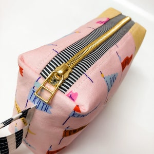 May include: A pink zippered pouch with a gold zipper and black and white striped trim. The pouch has a variety of colorful words printed on it, including "truth", "love", "kindness", and "compassion".