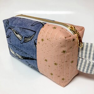 May include: A blue and pink zippered pouch with a whale print on one side and a gold star print on the other side. The pouch has a white and blue striped handle.