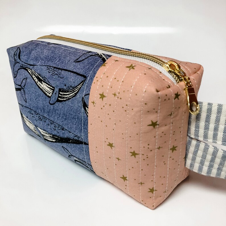 May include: A blue and pink zippered pouch with a whale print on one side and a gold star print on the other side. The pouch has a white and blue striped handle.