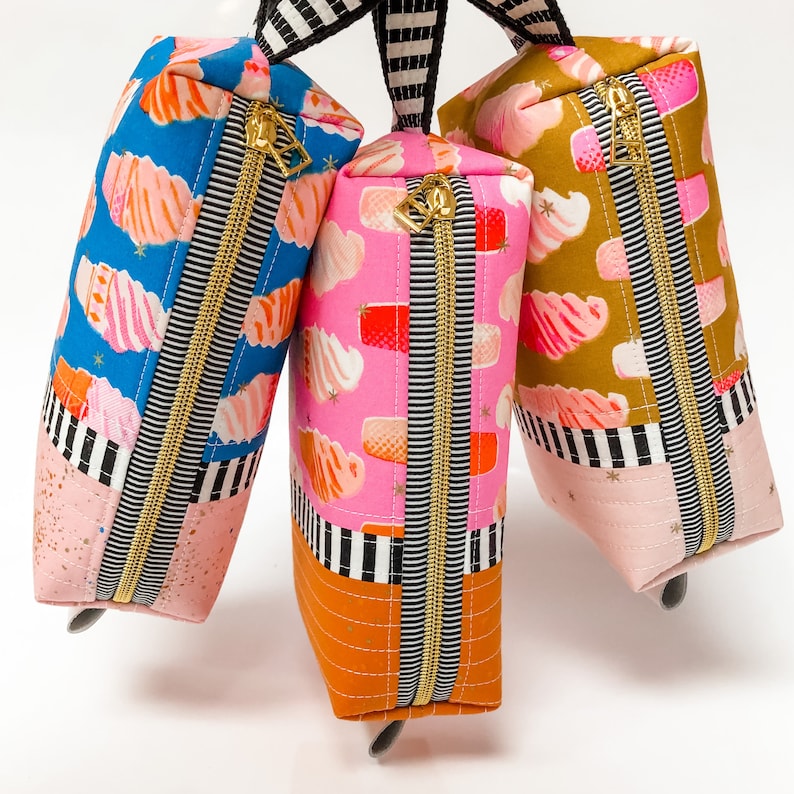 May include: Three zippered pouches with colorful patterned fabric. The pouches are pink, blue, and yellow with black and white stripes. Each pouch has a gold zipper.
