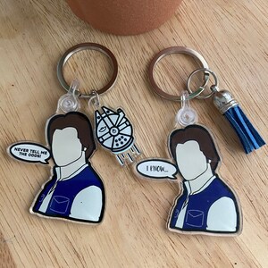 May include: Two clear acrylic keychains featuring a cartoon illustration of Han Solo from Star Wars. One keychain has the text "Never tell me the odds!" and the other keychain has the text "I know..."