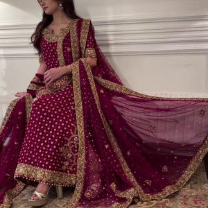 Könnte beinhalten: Ein formelles bordeauxfarbenes Ensemble mit goldener Stickerei. Das Outfit umfasst ein langärmliges Oberteil, eine passende Hose und einen transparenten Dupatta. Die aufwendigen goldenen Details zieren den Ausschnitt, die Ärmel und die Ränder und schaffen einen luxuriösen Look. Das Outfit wird mit goldenen Absätzen getragen.