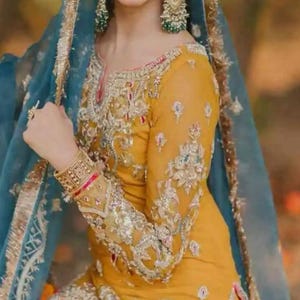 May include: A woman wearing a mustard yellow embroidered dress with a matching headpiece and earrings. The dress features intricate silver and gold embroidery. A sheer blue veil is draped over the head and shoulders. The overall look is elegant and festive.