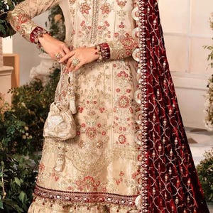 May include: An elegant cream-colored formal dress with intricate gold embroidery and red accents. The dress features long sleeves, a ruffled skirt, and a matching purse. A deep red velvet shawl with ornate detailing completes the ensemble.