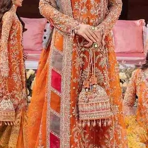 May include: A woman wearing a traditional orange and gold embroidered dress with a matching dupatta. The dress features intricate silver and gold embellishments and a red and pink border. The woman is holding a small, ornate clutch bag.