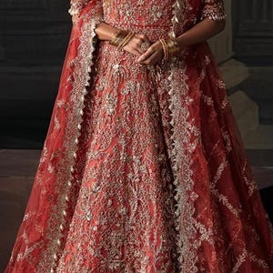 Op de afbeelding: Een rode formele jurk met ingewikkelde gouden borduursels en kralenwerk. De jurk heeft lange mouwen, een volle rok en een bijpassende sjaal. Gouden armbanden en een ketting completeren de jurk. De algehele stijl is elegant en sierlijk.