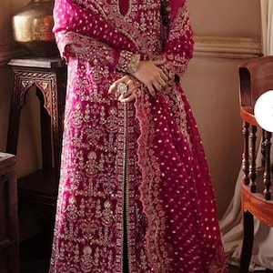 May include: A magenta-colored, embroidered formal dress with a matching shawl. The dress features intricate gold embellishments and a keyhole neckline. The long sleeves and floor-length design create an elegant silhouette, perfect for special occasions.