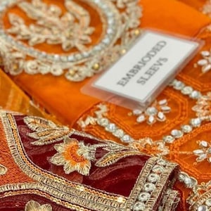 May include: Close-up of a maroon and orange embroidered fabric with gold trim and sequins. The fabric is folded and a white tag with the text "Embroidered Sleeves" is visible.