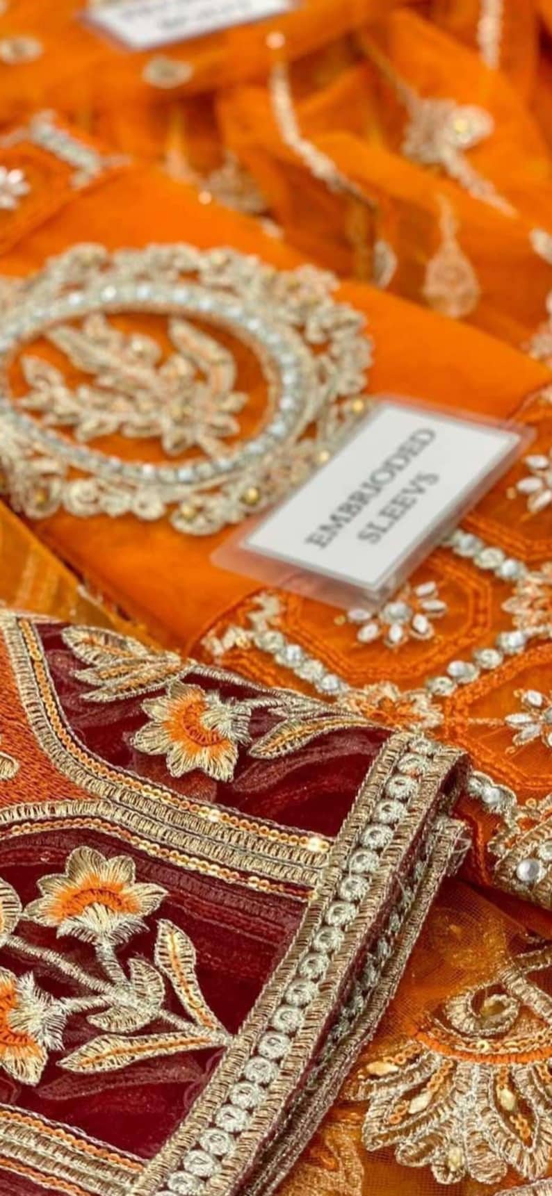 May include: Close-up of a maroon and orange embroidered fabric with gold trim and sequins. The fabric is folded and a white tag with the text "Embroidered Sleeves" is visible.