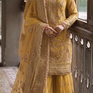 May include: Mustard yellow embroidered formal wear ensemble. The outfit includes a long-sleeved tunic, matching pants, and a sheer dupatta. The intricate gold embroidery and embellishments add a touch of elegance to the traditional attire.