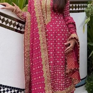 May include: A magenta-colored, long-sleeved dress with gold embroidery and a matching scarf. The dress features a detailed pattern and is worn with beige pants and silver heels. The outfit is suitable for special occasions.