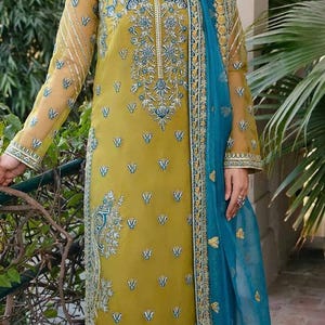 May include: Mustard yellow embroidered salwar kameez with a teal blue dupatta. The long-sleeved tunic features intricate floral embroidery in teal and silver. The matching pants and dupatta are adorned with similar detailing.