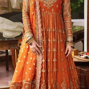 May include: A woman wearing a long, orange dress with intricate gold embroidery and a matching dupatta. The dress has a flared skirt and a fitted bodice. The woman is wearing gold jewelry, including a necklace, earrings, and bangles.