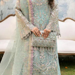 May include: A light blue embroidered formal dress with a matching clutch. The dress features intricate detailing, long sleeves, and a flowing skirt. The clutch is embellished with beads and tassels. The outfit is suitable for special occasions.