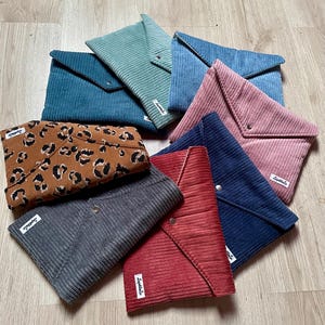 Può includere: Una collezione di pochette a busta in velluto a coste in vari colori, tra cui verde acqua, verde, rosa, blu navy, rosso e grigio. Una pochette presenta un motivo leopardato. Ogni pochette ha una chiusura a patta e una piccola etichetta.