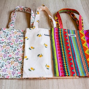 Peut inclure: Trois sacs fourre-tout avec différents motifs. L'un présente un motif floral avec des feuilles roses, violettes et vertes. Un autre a un motif de citron sur fond blanc. Le troisième a un motif géométrique coloré avec des rayures rouges, vertes et jaunes.