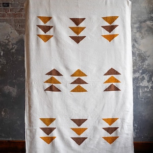 May include: White throw blanket with a geometric pattern of brown and gold triangles. The blanket has a fringed edge.
