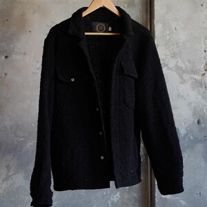 May include: A black textured jacket with two chest pockets and button closures, hanging on a wooden hanger. The jacket has a soft, fuzzy appearance and is displayed against a neutral gray background.