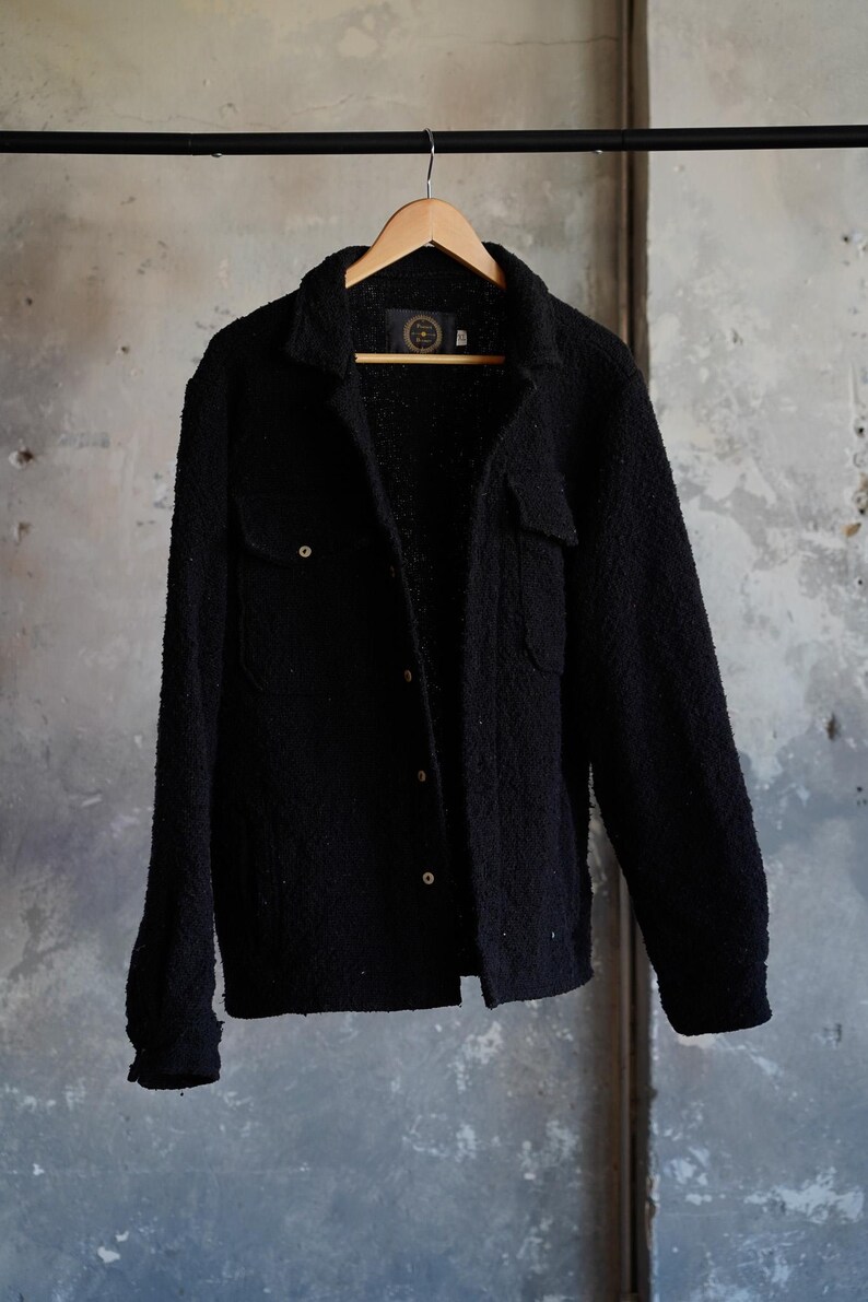 May include: A black textured jacket with two chest pockets and button closures, hanging on a wooden hanger. The jacket has a soft, fuzzy appearance and is displayed against a neutral gray background.