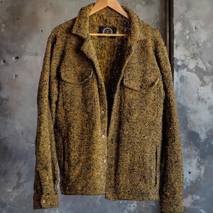 May include: A textured, button-down jacket in shades of gold and black hangs on a wooden hanger. The jacket features two chest pockets and a classic collar. The fabric has a speckled appearance, and the garment is displayed against a neutral background.