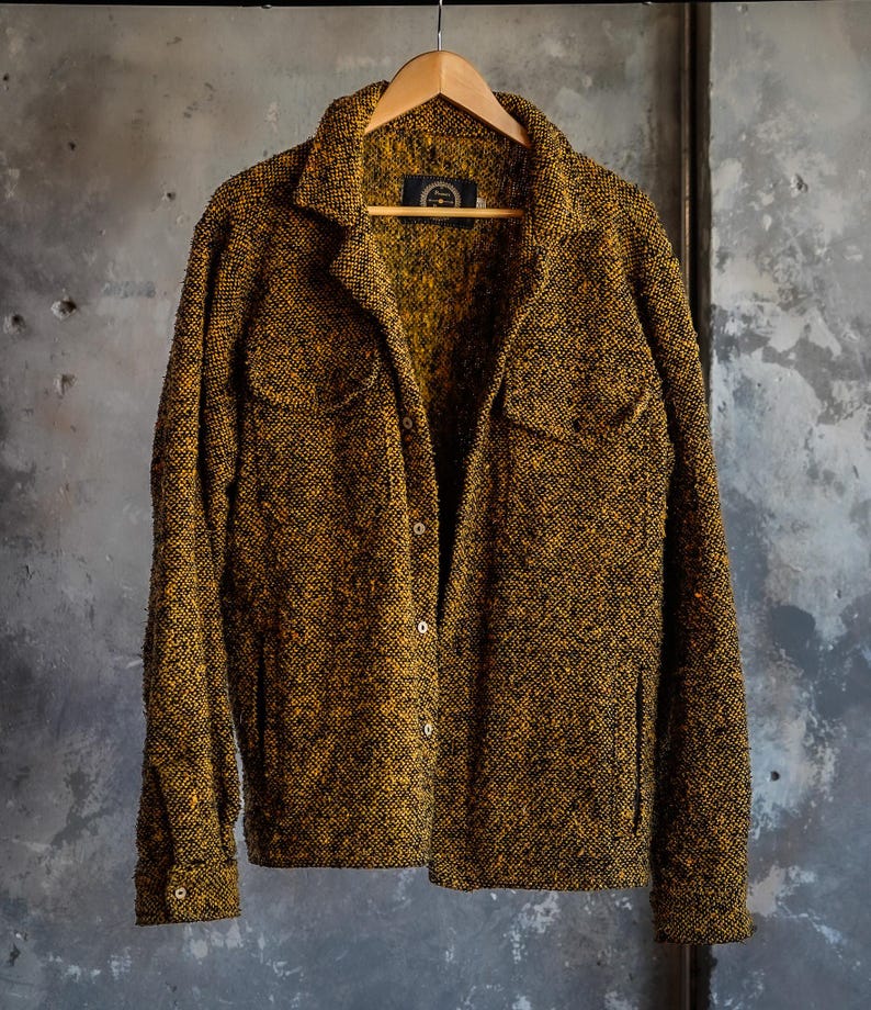 May include: A textured, button-down jacket in shades of gold and black hangs on a wooden hanger. The jacket features two chest pockets and a classic collar. The fabric has a speckled appearance, and the garment is displayed against a neutral background.