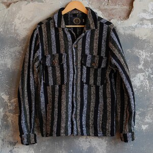 May include: A long-sleeved, button-down shirt with vertical stripes in black, green, and white. The shirt has two chest pockets and is hanging on a wooden hanger against a textured wall.