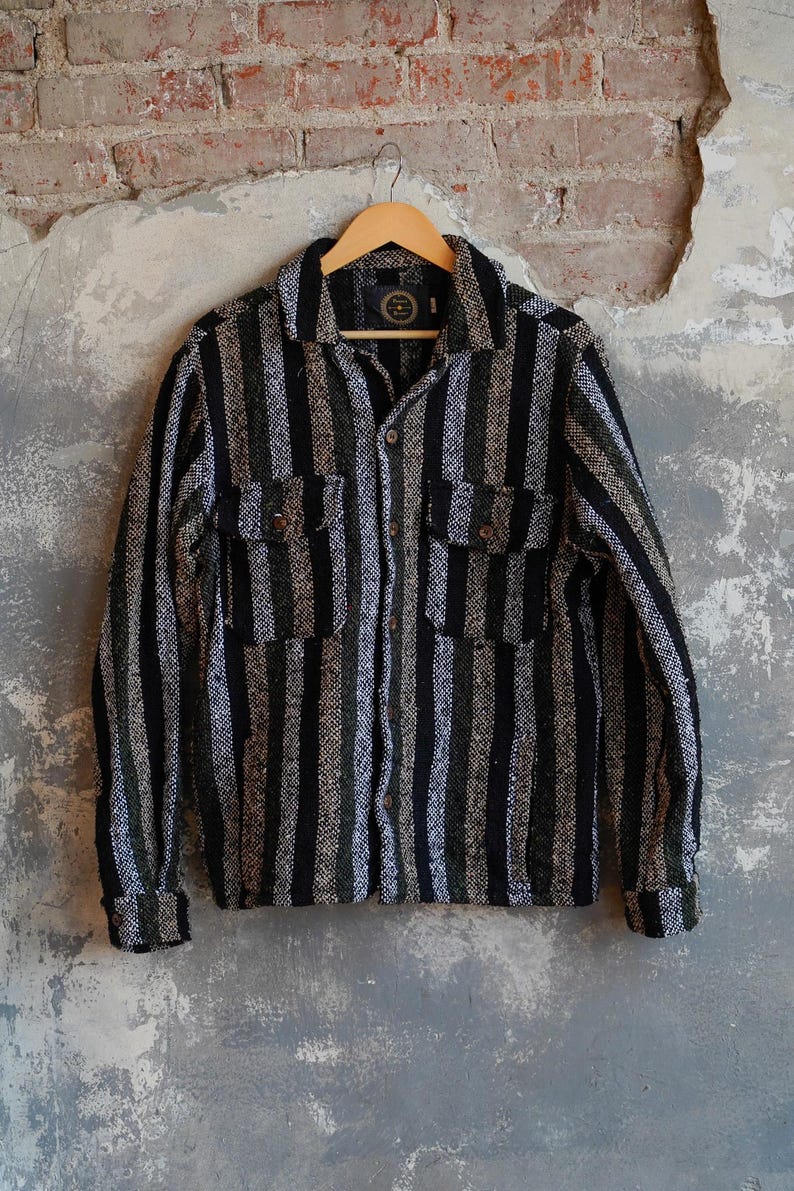 May include: A long-sleeved, button-down shirt with vertical stripes in black, green, and white. The shirt has two chest pockets and is hanging on a wooden hanger against a textured wall.