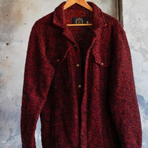May include: A red textured jacket with two chest pockets and button closures hangs on a wooden hanger. The jacket has a collared neckline and long sleeves. The background is a neutral, textured wall.