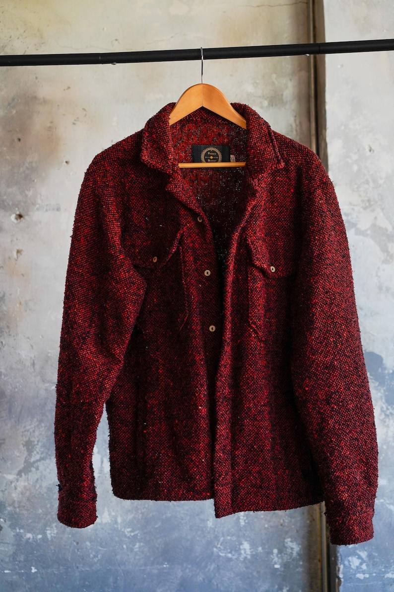 May include: A red textured jacket with two chest pockets and button closures hangs on a wooden hanger. The jacket has a collared neckline and long sleeves. The background is a neutral, textured wall.