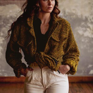 May include: A woman wearing a mustard-colored woven jacket with two front pockets and a black shirt. She is also wearing off-white pants. The jacket has a textured appearance. The background is a neutral color.
