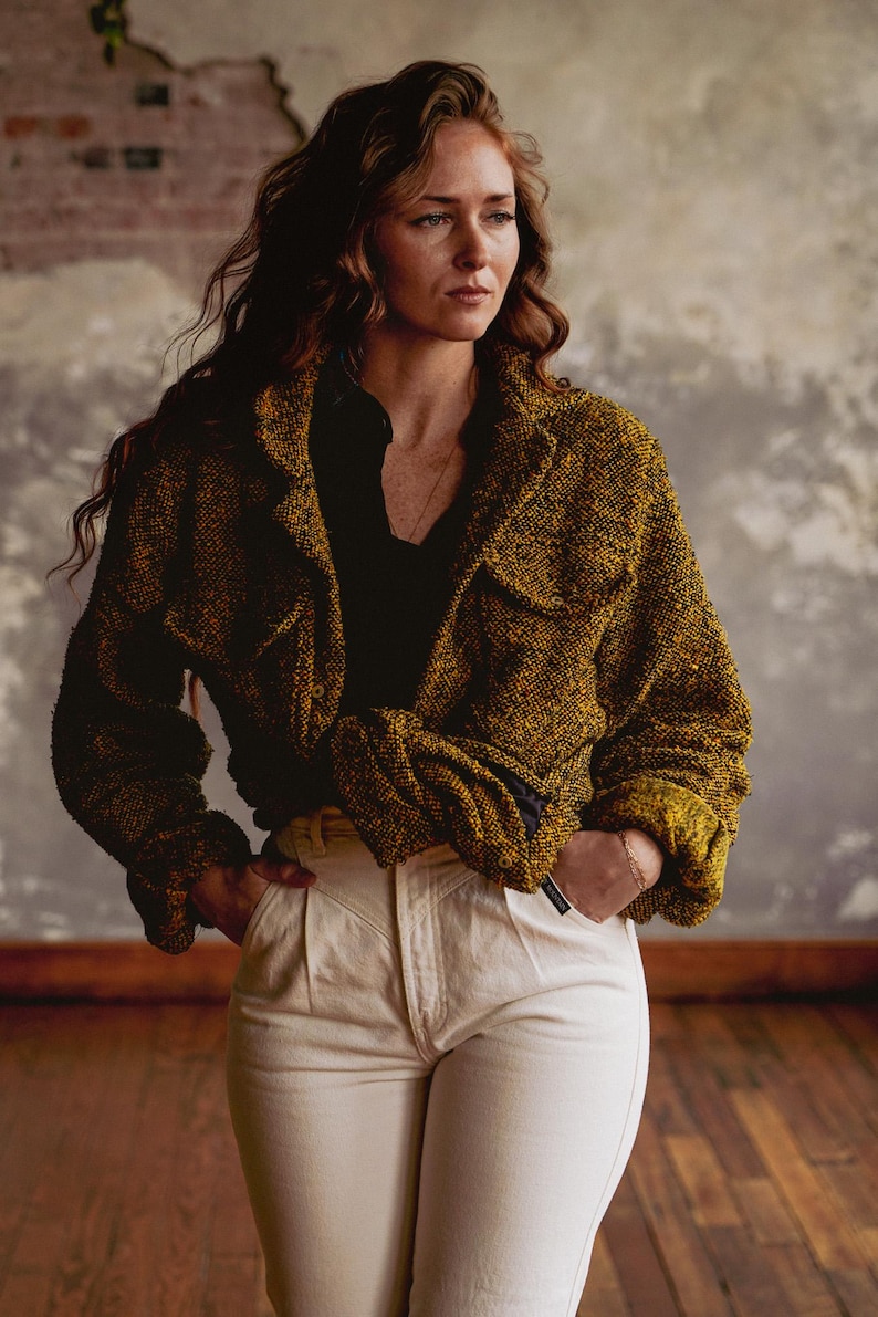 May include: A woman wearing a mustard-colored woven jacket with two front pockets and a black shirt. She is also wearing off-white pants. The jacket has a textured appearance. The background is a neutral color.