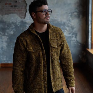 May include: A textured, button-down jacket in shades of olive green and gold. The jacket features two chest pockets and a classic collar. The person in the image is wearing a black t-shirt and dark jeans. The background is a textured wall and wooden floor.
