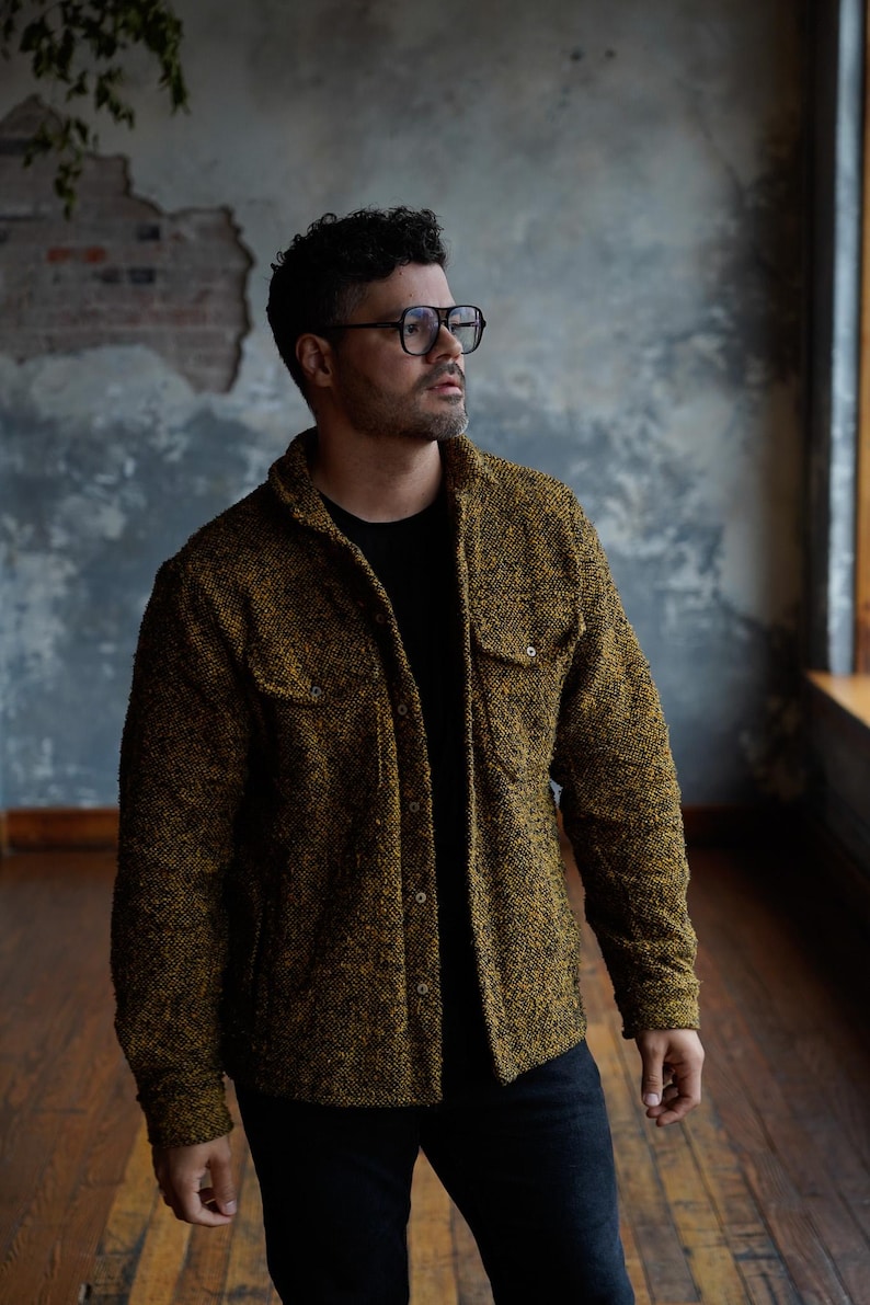 May include: A textured, button-down jacket in shades of olive green and gold. The jacket features two chest pockets and a classic collar. The person in the image is wearing a black t-shirt and dark jeans. The background is a textured wall and wooden floor.
