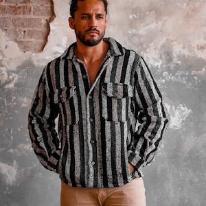 May include: A black and white striped button-down shirt with two chest pockets. The shirt is worn open, revealing the chest. The model is wearing tan pants. The background is a textured wall.