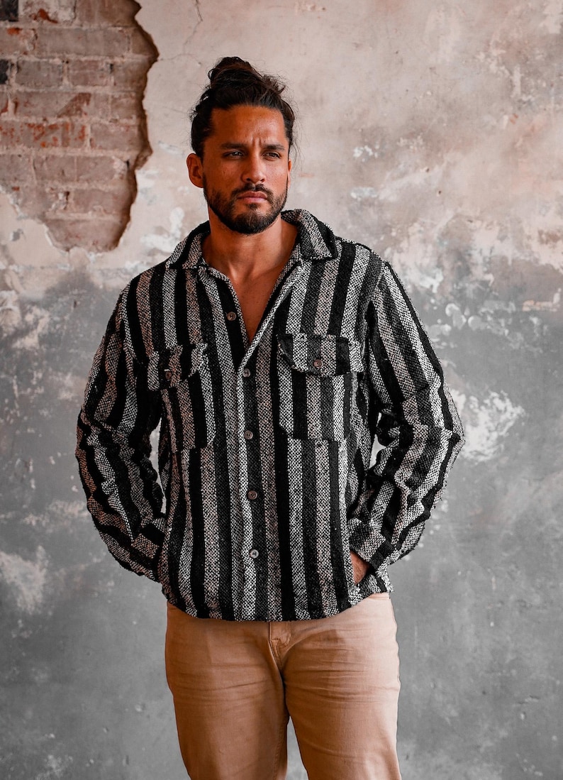 May include: A black and white striped button-down shirt with two chest pockets. The shirt is worn open, revealing the chest. The model is wearing tan pants. The background is a textured wall.