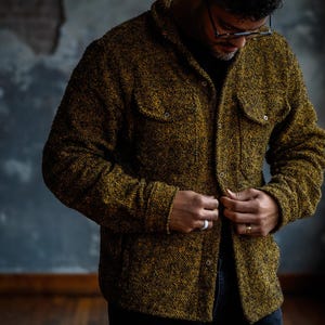 May include: A textured, button-down jacket in shades of gold and black. The jacket features two chest pockets and is being buttoned up. The person in the image is wearing black pants and glasses.
