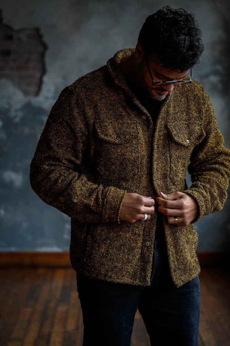 May include: A textured, button-down jacket in shades of gold and black. The jacket features two chest pockets and is being buttoned up. The person in the image is wearing black pants and glasses.