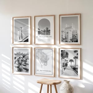 May include: Six framed black and white prints of St. Petersburg, Florida, with wooden frames. The prints depict a bridge, cityscapes, a map, and a boardwalk with palm trees. A wooden stool and a decorative vase are in the foreground.