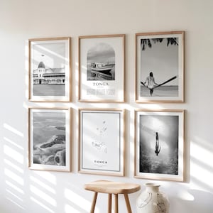 May include: Six framed black and white photographs with wooden frames are displayed on a white wall. The images depict various scenes, including a building, a boat, a person on a hammock, aerial views, a map, and a diver. The word "TONGA" is visible on two of the images.