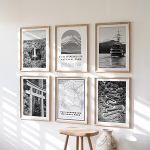 Puede incluir: Seis impresiones en blanco y negro enmarcadas con marcos de madera se exhiben en una pared blanca. Las impresiones representan paisajes, un barco y un mapa. Una impresión presenta el texto "FUJI-HAKONE-IZU NATIONAL PARK". Un taburete de madera y un jarrón decorativo están frente a las impresiones.