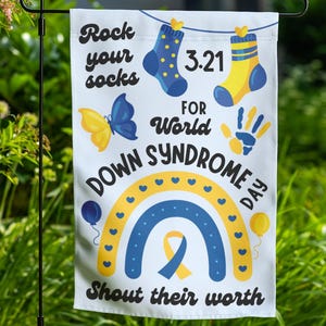 K&ouml;nnte beinhalten: Eine wei&szlig;e Gartenflagge mit einem blauen und gelben Regenbogen, einem gelben Band und dem Text "Rock your socks for World Down Syndrome Day. Shout their worth." Die Flagge zeigt au&szlig;erdem einen Schmetterling, einen Handabdruck und zwei Socken.
