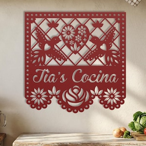 Op de afbeelding: Rode metalen wanddecoratie met een decoratief ontwerp. Het ontwerp omvat twee vogels, een hart met bloemendetails en de tekst "Tia's Cocina". De onderste rand heeft een geschulpte bloemenpatroon.