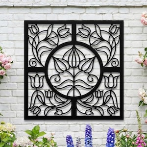 Op de afbeelding: Een zwart metalen wandkunstwerk met een bloemenontwerp. Het vierkante kunstwerk heeft een centraal cirkelvormig motief omgeven door gestileerde bloemen en bladeren, tegen een witte bakstenen muur en omgeven door kleurrijke bloemen.