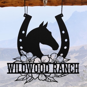 May include: Black metal Wildwood Ranch sign featuring a horse head silhouette inside a horseshoe, adorned with floral accents. The sign is suspended by chains from a wooden beam, with a skull and horns above.