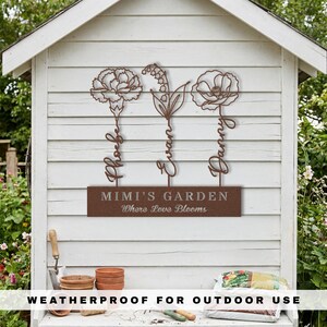 K&ouml;nnte beinhalten: Ein braunes Metall-Gartenschild mit drei stilisierten Blumendesigns und den Namen: Phoebe, Evan und Danny. Das Schild lautet "MIMI'S GARDEN Where Love Blooms" und ist mit "WEATHERPROOF FOR OUTDOOR USE" beschriftet. Das Schild ist an einer wei&szlig;en Holzkonstruktion befestigt.