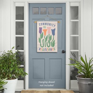 May include: A beige canvas flag with the text "COMMUNITY IS RESISTANCE" in colourful letters. The flag features a stylised design of four tulips in purple, orange, pink, and blue. The flag is hanging on a blue door with a wooden dowel.
