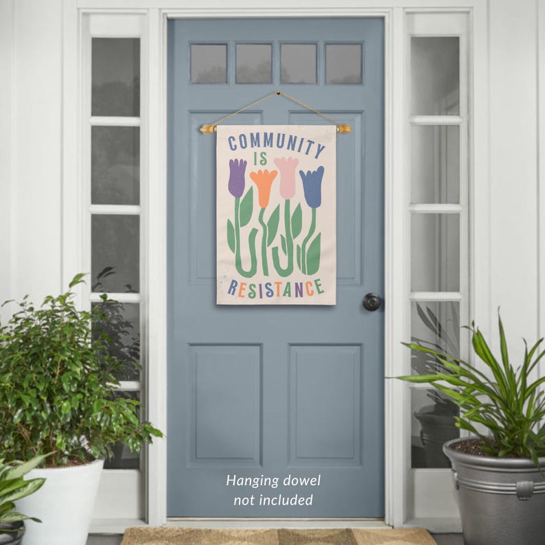 May include: A beige canvas flag with the text "COMMUNITY IS RESISTANCE" in colourful letters. The flag features a stylised design of four tulips in purple, orange, pink, and blue. The flag is hanging on a blue door with a wooden dowel.