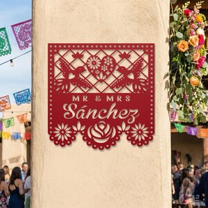 Op de afbeelding: Een rood metalen bord met de woorden "MR & MRS S&aacute;nchez" uitgesneden in een decoratief lettertype. Het bord heeft een hart, vogels en bloemmotieven, ideaal voor een bruiloft of jubileum.