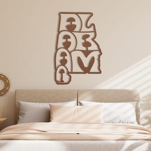Puede incluir: Adorno de pared de madera con la palabra "ALABAMA" escrita en una fuente apilada y estilizada. Las letras son de color marr&oacute;n c&aacute;lido, con un contorno marr&oacute;n m&aacute;s claro. La obra de arte est&aacute; montada en una pared de color neutro sobre una cama.